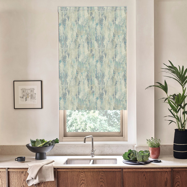 Patterned roller blind covers a window above a sink; faucet centered on a marble countertop with wooden cabinets, bowl of leafy greens, potted plants and framed sketch on wall.