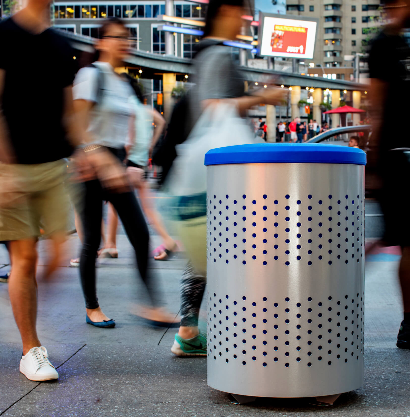 silver powder coated steel recycling bin with a blue liner and blue full opening lid outdoors on a busy city sidewalk with people walking by