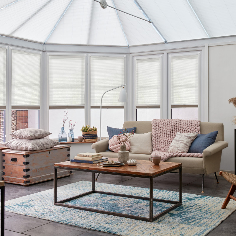 White pleated cellular shades span the bay windows and conservatory roof, lowered to diffuse daylight; cosy living room with beige sofa, chunky pink throw, wooden coffee table and blue rug.