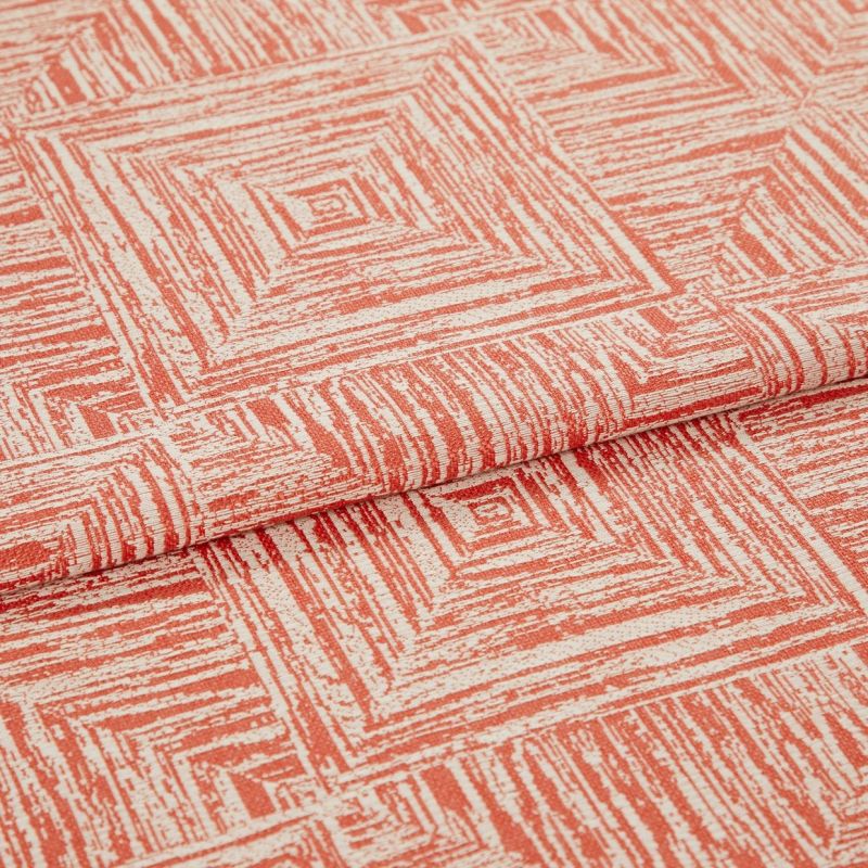 Folded coral-and-cream fabric with textured concentric-square print; a layered fold reveals repeating pattern, shown close-up on a flat surface under soft, even lighting.