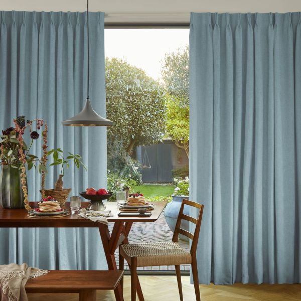 Floor-length pleated pale-blue curtains, drawn slightly apart to frame a central opening, revealing a sunlit dining table set for breakfast and a lush backyard garden beyond.