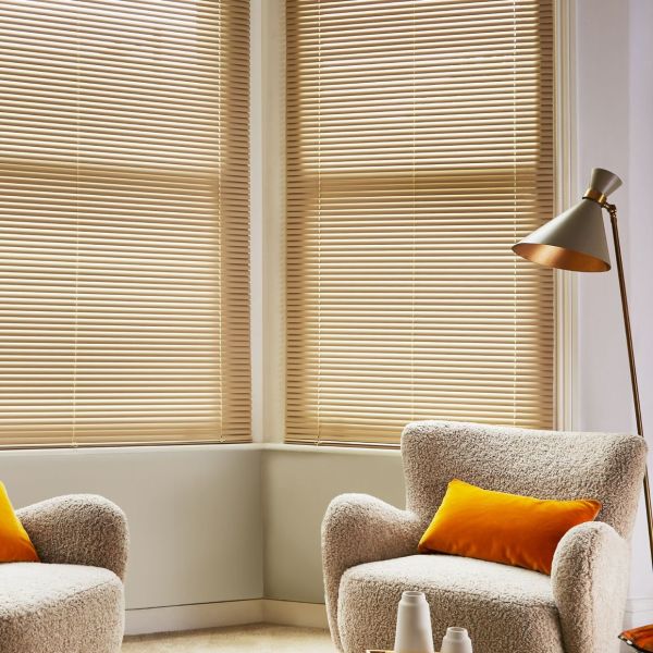 Horizontal beige venetian blinds covering bay windows, tilted closed to softly filter warm daylight; in a cozy living-room corner with textured boucle armchairs, orange cushions, a floor lamp, and neutral walls.