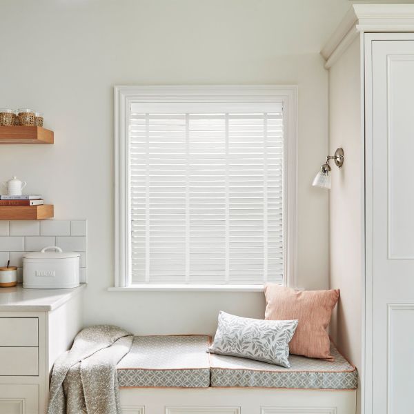 White horizontal venetian blinds (slatted, closed) covering the window, filtering soft daylight; bright neutral kitchen nook with cushioned bench, throw, wall sconce, countertop and bread box.
BREAD
THE PIG