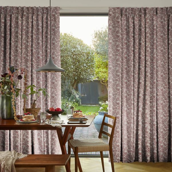 Pleated floor-length curtains with red leaf pattern, parted slightly, filtering daylight through a glass door; breakfast table, pendant lamp and wooden chairs in a dining room opening onto a garden.