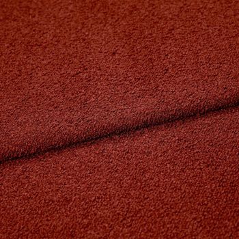 Rust-red textured boucle fabric folded across the frame, creating a dark seam; close-up studio view highlighting coarse looped surface and even, warm lighting.