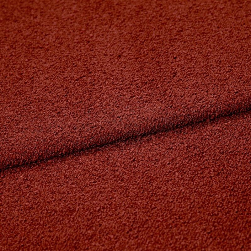 Rust-red textured boucle fabric folded across the frame, creating a dark seam; close-up studio view highlighting coarse looped surface and even, warm lighting.