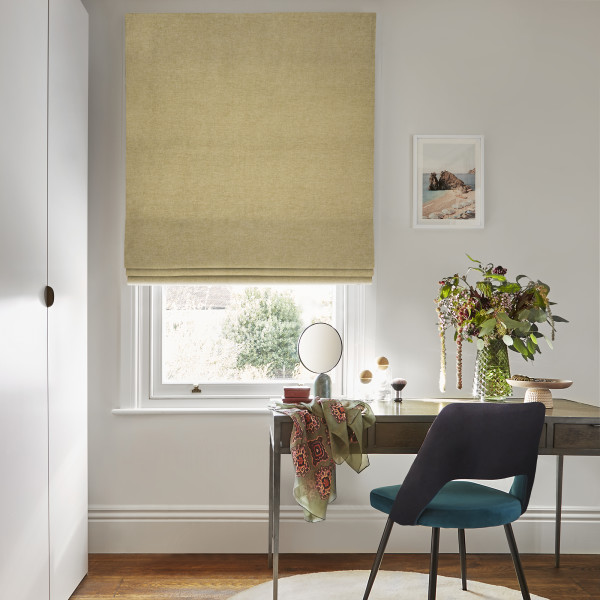 Beige Roman blind, lowered with soft folds and textured fabric, filtering daylight through the window; home workspace beneath with a desk, teal chair, vase of flowers and framed seaside print.