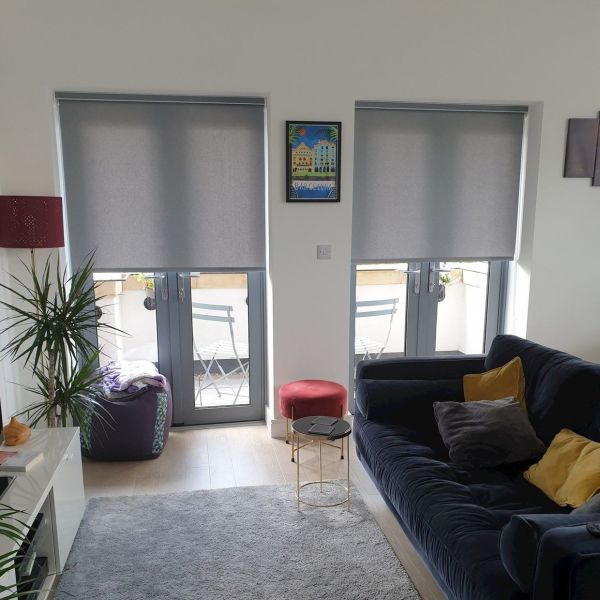 Two grey roller blinds lowered over adjacent French doors, filtering daylight and leaving bottom panes exposed; living room with dark sofa and yellow cushions, side table, plant, balcony chairs.
