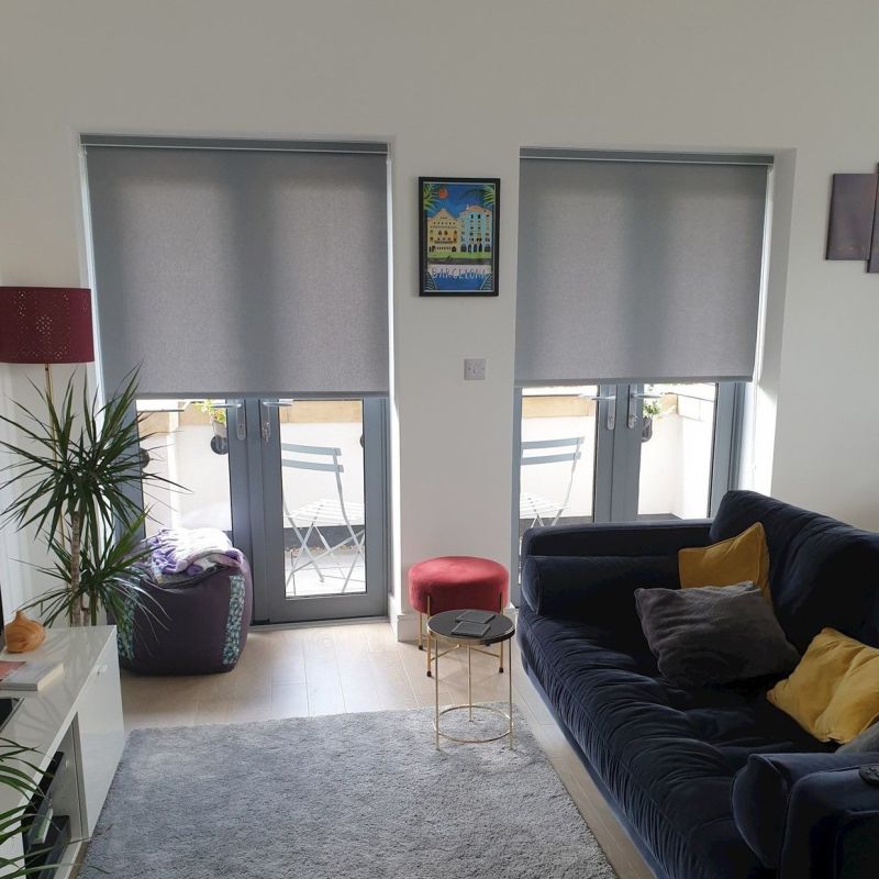 Two grey roller blinds lowered over adjacent French doors, filtering daylight and leaving bottom panes exposed; living room with dark sofa and yellow cushions, side table, plant, balcony chairs.