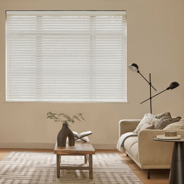 White horizontal Venetian blinds covering a large window, fully closed and filtering daylight; beige living room with sofa, wooden coffee table, patterned rug and slim black floor lamp.