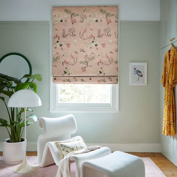 Pink Roman blind with crane motifs, partially lowered, filtering daylight into a pale-green sitting room with lounge chair, floor lamp, potted plant, framed bird print and hanging robe.