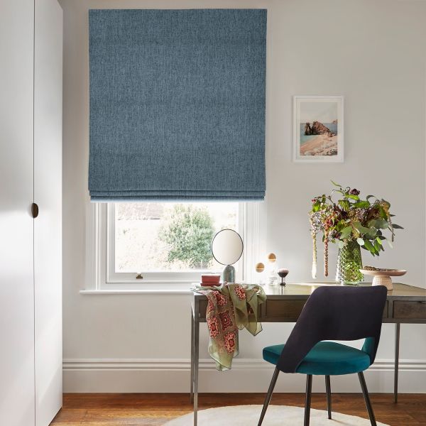 Blue textured Roman blind lowered over a sash window, filtering daylight; a wooden desk beneath holds a vase of flowers, small mirror and teal-upholstered chair in a neutral, sunlit room.
