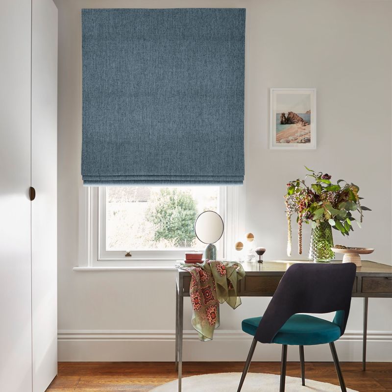 Blue textured Roman blind lowered over a sash window, filtering daylight; a wooden desk beneath holds a vase of flowers, small mirror and teal-upholstered chair in a neutral, sunlit room.