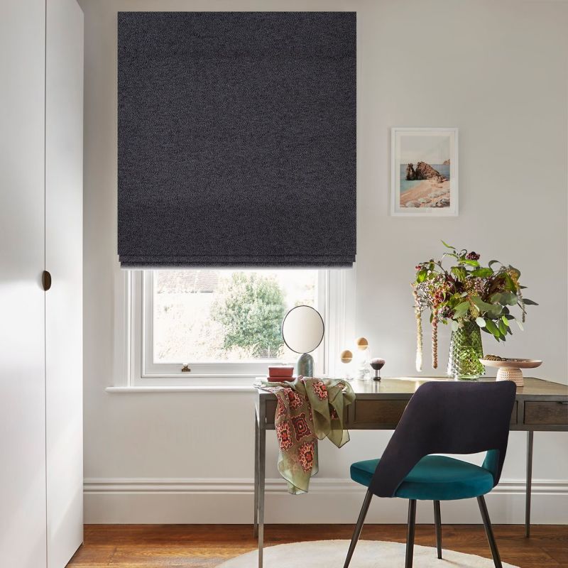Dark grey Roman blind lowered over a sash window, nearly closed, raised at the hem and filtering daylight; home desk with teal chair, tabletop mirror, vase of flowers and a draped scarf.