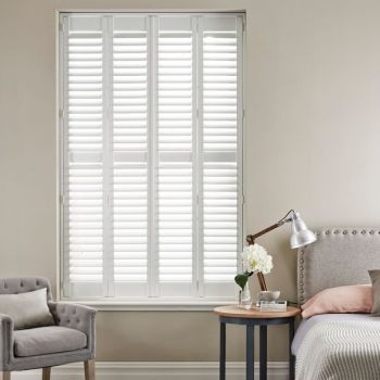 White plantation shutters covering a floor-to-ceiling window; slatted panels closed, filtering soft daylight. Context: neutral-toned bedroom nook with upholstered armchair, bedside table, lamp and floral vase.