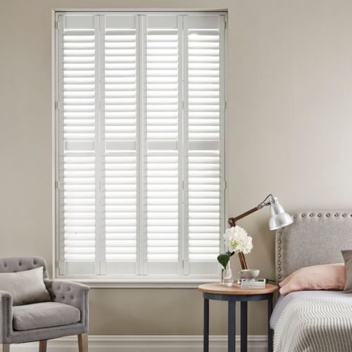White plantation shutters covering a floor-to-ceiling window; slatted panels closed, filtering soft daylight. Context: neutral-toned bedroom nook with upholstered armchair, bedside table, lamp and floral vase.