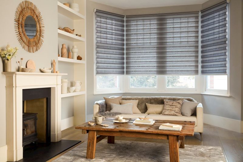 Grey layered zebra roller blinds covering three bay-window panels, partially lowered and filtering soft daylight; modern living room with beige sofa, rustic wooden coffee table, fireplace and neutral decor.