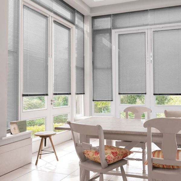 Horizontal aluminium Venetian blinds on tall French windows and doors, lowered with slats slightly tilted to filter daylight; sunlit dining nook with wooden table and chairs, patterned cushions, garden view.