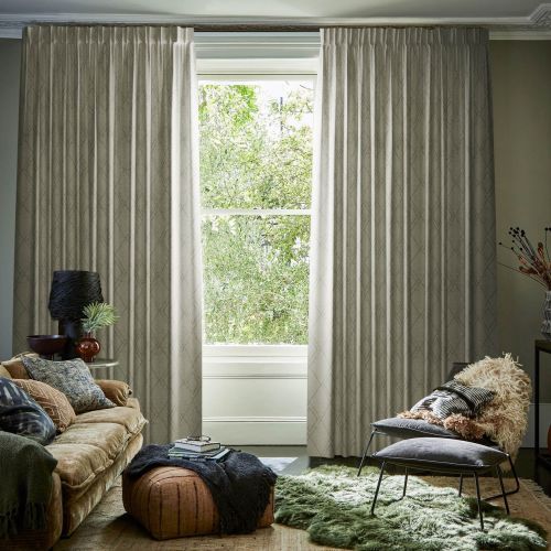 Heavy pleated beige curtains hang from a ceiling rod, parted centrally to reveal a bright window; living room with plush sofa, ottoman, shaggy green rug and leafy outdoor view.