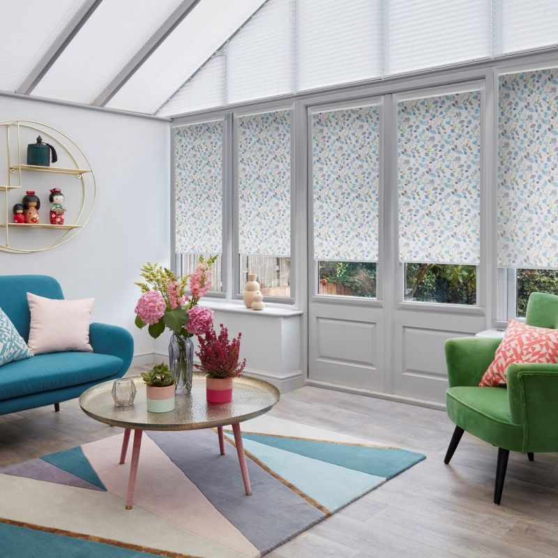 Floral-patterned roller blinds, lowered across tall conservatory windows and doors with a roof blind above, filtering soft daylight; bright sunroom with teal sofa, green armchair, coffee table and garden view.