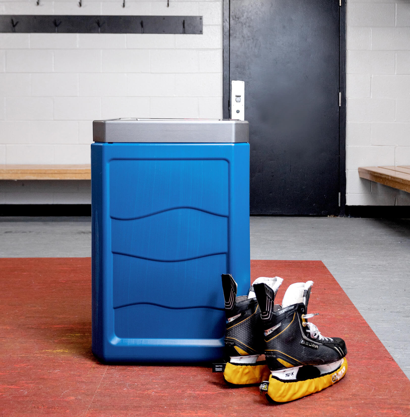 blue plastic recycling container with a brushed pewter plastic lid inside a hockey change room with a pair of skates on the floor beside the bin