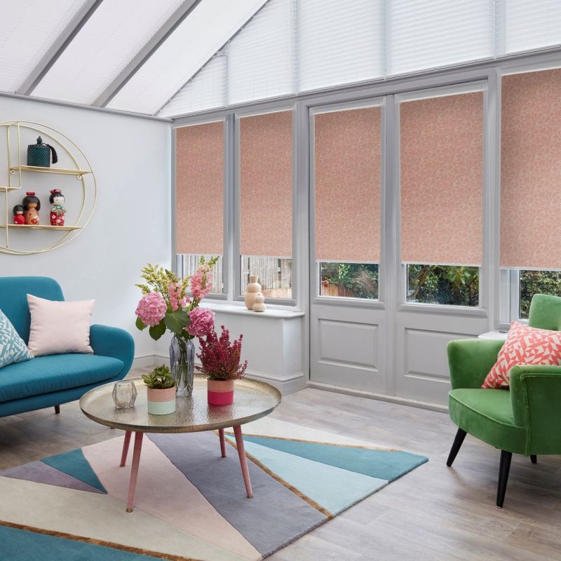 Patterned pink roller blinds, lowered across four tall windows and glass door, swirled print; filtering daylight and obscuring most view; bright conservatory with slanted glass roof, colorful seating and flowers.