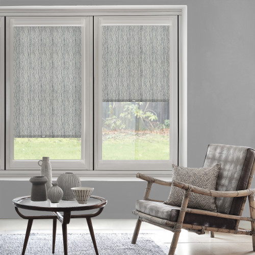 Roller blinds with a subtle vertical wave pattern, left fully lowered and right halfway, filtering daylight; in a grey-walled living room with a rattan armchair, coffee table and lawn view.