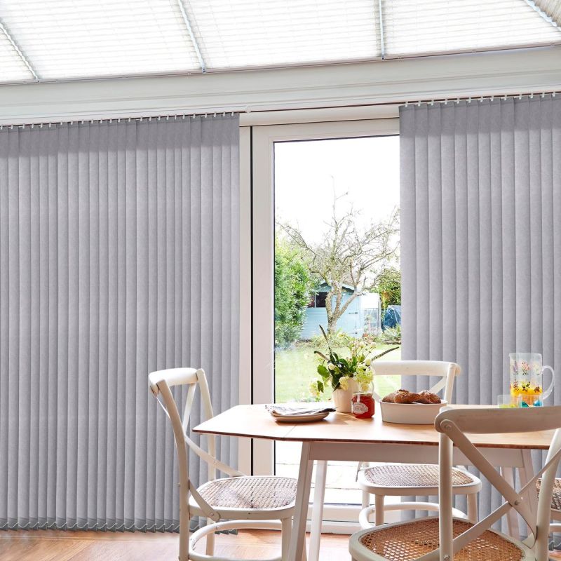 Grey vertical blinds on a track, full-height slatted panels parted at the center over a glass sliding door, partially open filtering daylight; small dining table, chairs, vase, garden outside.