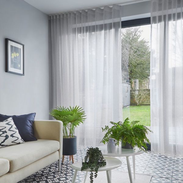 Sheer floor-to-ceiling grey curtains with soft pleats, drawn across a sliding glass door, gently filtering daylight; living room with sofa, patterned tile floor, potted plants, and a grassy backyard.