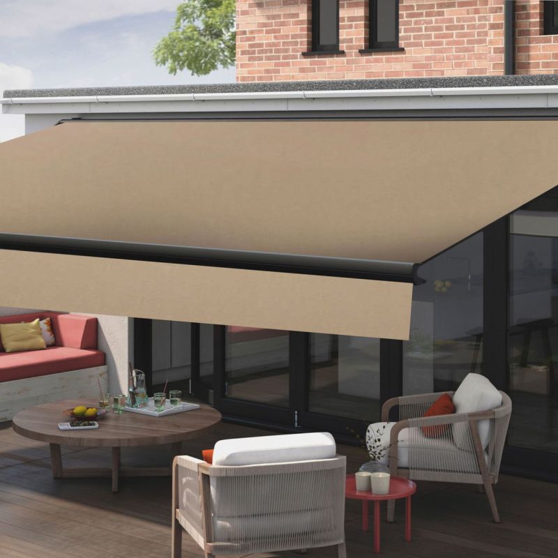 Beige retractable patio awning mounted above sliding glass doors, extended, casting shade over a wooden deck; modern outdoor seating, low round table with drinks, brick upper wall and tree.
