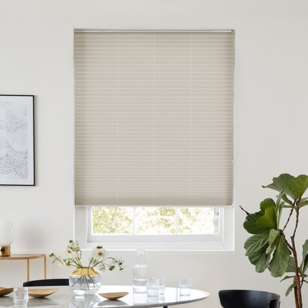 Beige pleated cellular blind lowered, with horizontal ribs and faint vertical cord lines, filtering soft daylight; over a bright minimalist dining nook with round table, vase, glassware and fiddle‑leaf fig.