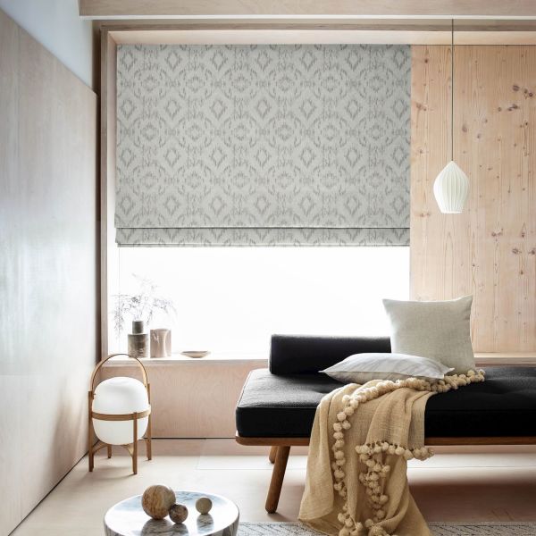 Patterned grey Roman blind, lowered with ikat motif, filters bright daylight over a window seat in a minimalist room with pale wood panels, a pendant light, cushions and a throw.