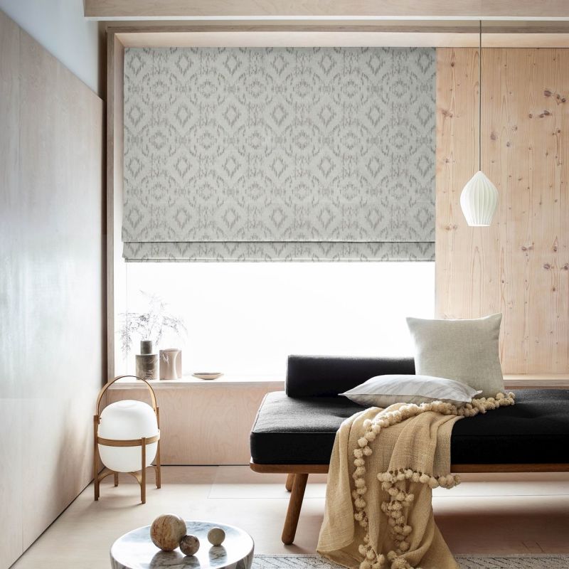 Patterned grey Roman blind, lowered with ikat motif, filters bright daylight over a window seat in a minimalist room with pale wood panels, a pendant light, cushions and a throw.