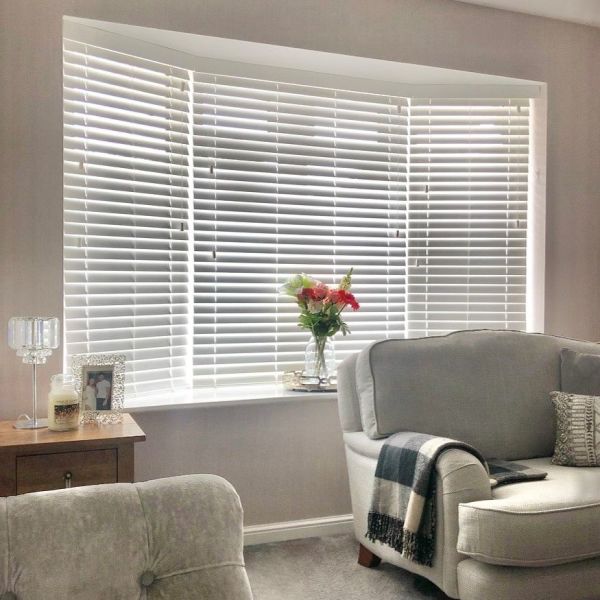 White horizontal Venetian blinds across a three-panel bay window, slats partially tilted letting soft daylight filter in; living room with gray upholstered armchairs, side table, vase of flowers on the sill.
