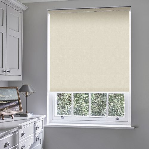 Beige fabric roller blind, fully lowered, subtly textured and light-filtering; covering a white sash window in a softly lit grey room with white cabinetry, dresser, lamp and leafy garden view.