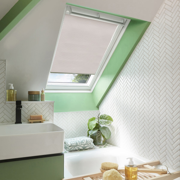 Pleated pale-pink skylight blind, mounted inside an angled roof window, partially lowered and filtering daylight; bathroom with mint-green trim, white herringbone tiles, sink, bathtub, plant, toiletries.