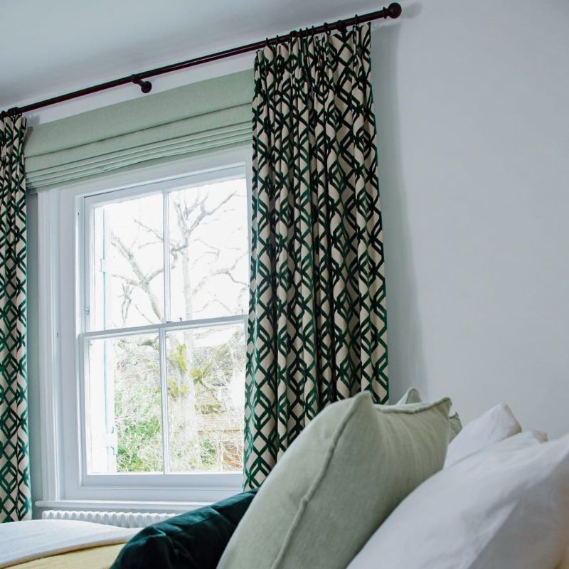 Green geometric pleated curtains hang on a dark rod, framing a lowered pale-green Roman blind that filters daylight; bedroom with pillows and a bare-tree view outside.
