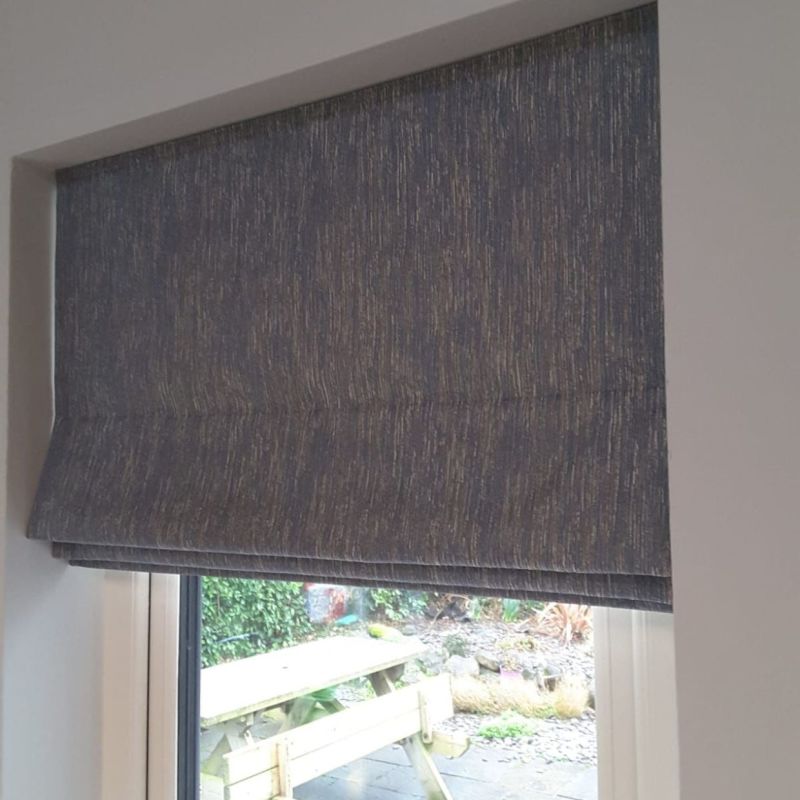 Grey textured Roman blind, lowered with visible horizontal folds, covers a recessed window, filtering daylight; interior white frame, partial garden view showing a wooden picnic bench outside.
