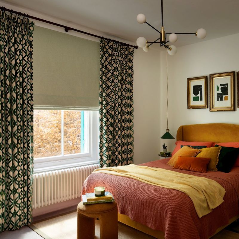 Patterned green-and-cream pleated curtains with a lowered pale Roman blind framing a sash window, filtering soft daylight into a cozy bedroom with mustard headboard, warm bedding, and pendant light.