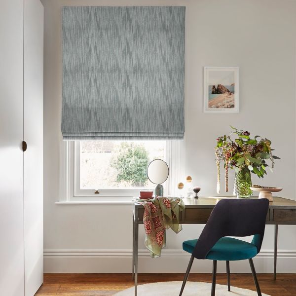 A textured grey Roman blind covers a window, lowered to filter daylight; a small desk with mirror, chair, vase of flowers and framed seaside print in a bright home office.