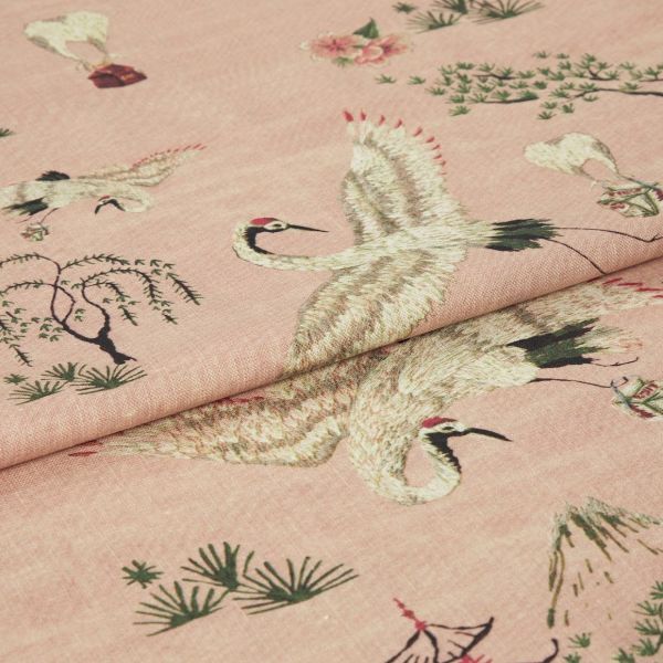 Pink woven fabric printed with red-crowned cranes and stylized trees — folded to show layered panels — close-up on a soft textile surface under soft, even lighting.