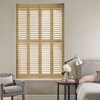 Wooden full-height plantation shutters covering tall window, divided into four louvered panels with tilted slats; closed, they softly filter daylight into a neutral bedroom with bed, side table, armchair.