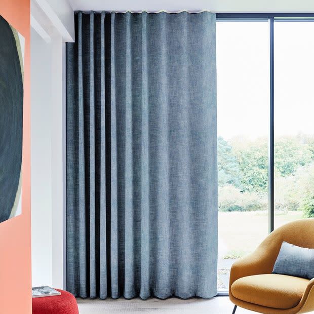 Floor-to-ceiling textured gray curtains with wave-top heading, mostly closed and filtering daylight from a large glass window; modern living room with mustard armchair and garden view.