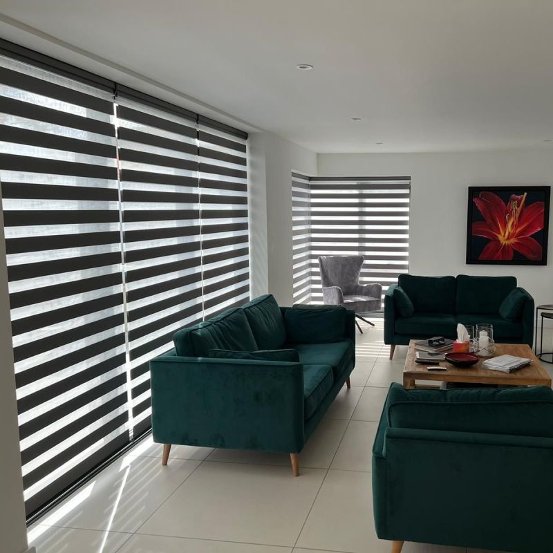 Horizontal zebra blinds on large corner windows, alternating opaque and sheer bands, filtering soft daylight into a modern living room with teal sofas, wooden coffee table, and a gray armchair.