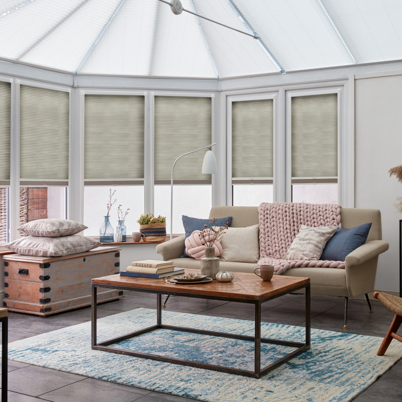 Beige pleated cellular blinds across six tall conservatory windows, lowered about three-quarters and filtering soft daylight; cozy sunroom with pitched translucent roof, neutral sofa, chunky pink throw, wooden coffee table.