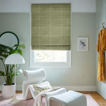 Patterned olive-green Roman blind, lowered with repeating geometric lattice; filtering daylight into a green sitting-room corner with white lounge chair, floor lamp, potted plant, framed bird print and yellow robe.