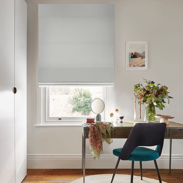 Light-gray Roman blind covering the window, lowered almost fully with a small gap; filtering soft daylight. In a neutral home office: wooden desk with mirror, vase of flowers, teal chair.