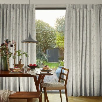 Full-length pleated pale-gray curtains with subtle floral sprigs, parted at center to reveal the glass door and filter soft daylight; dining table set for breakfast in a sunlit garden-facing room.