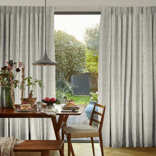 Full-length pleated pale-gray curtains with subtle floral sprigs, parted at center to reveal the glass door and filter soft daylight; dining table set for breakfast in a sunlit garden-facing room.