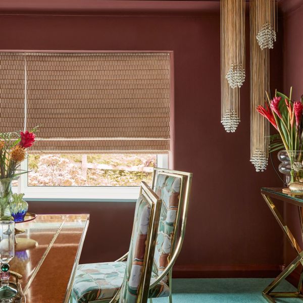 Woven beige patterned roman blind lowered over a window, filtering daylight with its bottom slightly raised; in a burgundy dining room with brass-framed floral chairs, wooden table, and beaded pendant lights.